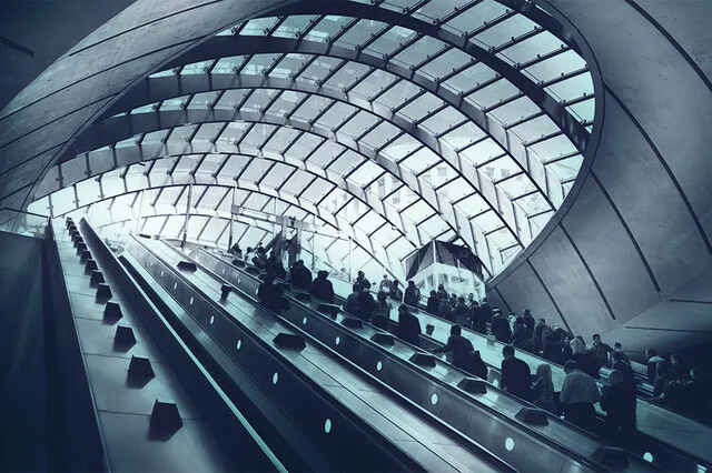The image shows a number of long escalators transporting crowds of people upwards from an underground station. The roof and building structure is of a futuristic design made of lots of metal and glass.