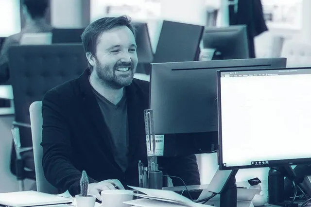 An image of a male sat at a desk and smiling at a computer screen. Situated around him are more desks with chairs, screens, and stationary.