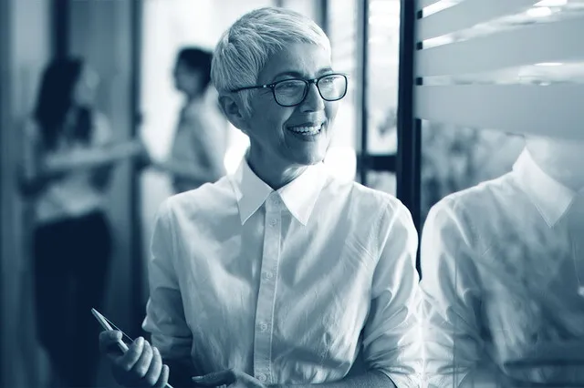 A photographic image showing a woman with short hair and glasses. She is smiling while holding a tablet and looking out of the window. There are two people chatting in the blurred background.