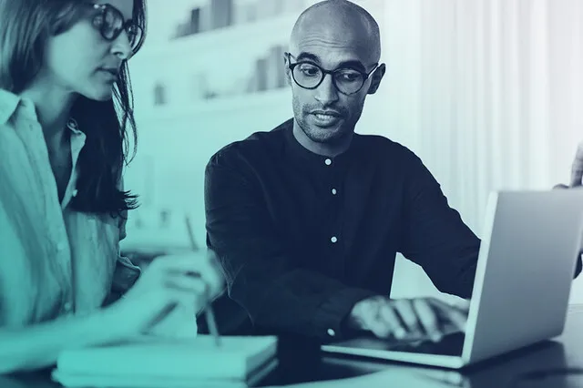 A photographic image showing a man and woman working together at a desk. The man is wearing glasses and explaining something on a laptop to the woman, who is taking notes with a pen and notepad.