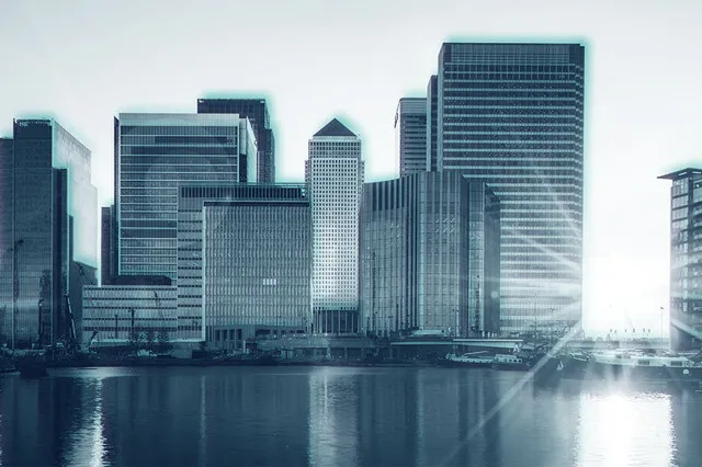 The image shows a city view of tall, modern skyscrapers situated along a waterfront. A low sunlight is bright in the right hand side of the image, bursting through between two buildings.
